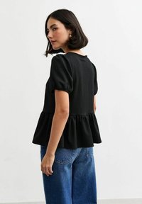 Svart texturerad blus med puffärmar och peplumslut, i kombination med lösa blå denimbyxor. Neutral bakgrund.