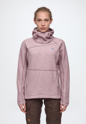 Une femme présente un pull en polaire mauve à col montant drapé et un pantalon marron, sur un fond clair uni.