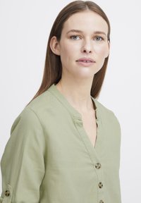Blouse verte claire à manches boutonnées avec un col en V, présentant un tissu texturé et un ourlet arrondi. Comprend des manches repliables avec des boutons en option.