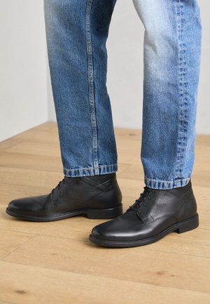 Stivali alla caviglia in pelle nera con una finitura liscia, punta tonda e design con lacci, abbinati a jeans in denim azzurro chiaro, su un pavimento in legno.