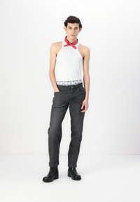Top senza maniche bianco, jeans neri e stivaletti neri alla caviglia. Bandana rossa attorno al collo. La modella è in piedi di fronte a uno sfondo bianco semplice.