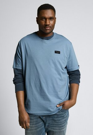 Jonge man met kort haar staat frontaal, draagt gelaagde blauwe overhemden en spijkerbroek, met één hand in zijn zak tegen een effen achtergrond.