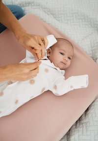 Vit babyklädsel med bruna solmönster, som kläs av en hand. I bakgrunden finns en rosa skötpad och en strukturerad grå filt.