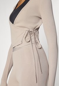 Bluzka z beżowej prążkowanej tkaniny z dekoltem w kształcie litery V. Posiada ozdobę w postaci paska w talii i długie rękawy, ułożona na ciemnym topie na ramiączkach.