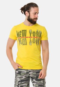 Gelbes Baumwoll-T-Shirt mit grafischem Aufdruck "NEW YORK", kombiniert mit grauen Camo-Cargo-Shorts mit Seitentaschen.