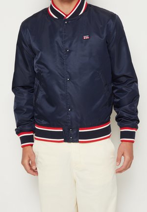Blouson bomber bleu marine avec col, poignets et ourlet côtelés rayés rouge, blanc et bleu marine, porté avec un pantalon beige clair.