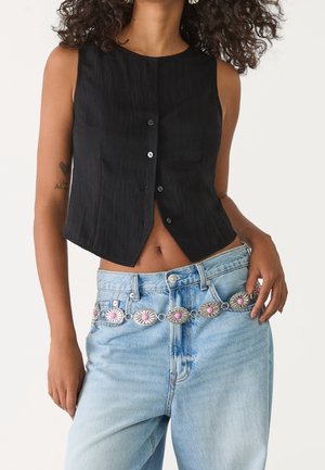 Kvinde iført en sort ærmeløs kort top med knapper og lyseblå jeans med et dekorativt sølv- og lyserødt concho-bælte.