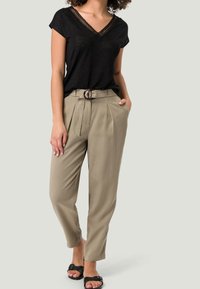 Svart V-ringad t-shirt med spetsdetaljer, ihopparad med beige högmidjade byxor med bälte och veck; modell som bär svarta slides.