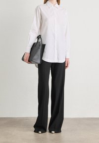 Persona che indossa una camicia bianca con bottoni, pantaloni larghi neri, scarpe bianche, mentre tiene una borsa nera in tessuto testurizzato su uno sfondo semplice.