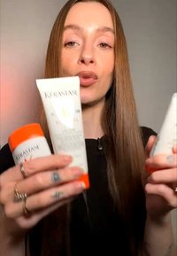 Mujer con cabello largo y castaño sosteniendo tres tubos de productos para el cabello Kérastase, dos con tapas naranjas, sobre un fondo liso.