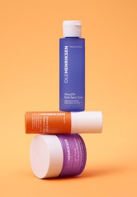 Trois produits de soin de la peau empilés : une bouteille de tonique violette, un tube de sérum orange et un pot de crème violet, avec des couvercles et des étiquettes blanches.