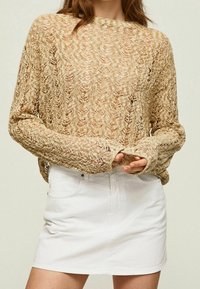 Pull beige en maille ouverte à manches longues, avec un motif texturé. Associé à une mini-jupe en denim blanche, offrant un look décontracté.