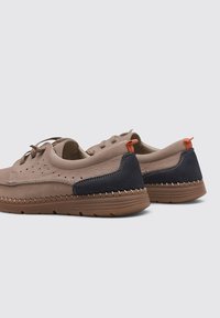 Chaussures en daim brun clair avec des accents bleu marine, design perforé, tirette orange et semelle en caoutchouc texturée. Minimalistes et fonctionnelles.
