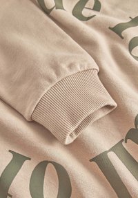 Felpa leggera beige con polsini a costina e grande scritta in verde scuro. Il tessuto sembra morbido con una texture liscia.