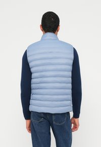 Világoskék párnázott mellény, vízszintes steppelt mintával, magas gallérral és cipzáras záródással, sötétkék hosszú ujjú ing fölött viselve.