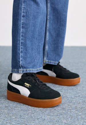 Sorte og hvide sneakers med suedes overflade og gummisul. Med guldbranding på siden og runde snørebånd, båret med blåt denim.
