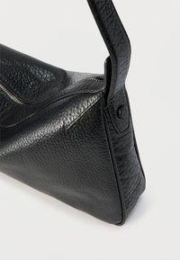 Fekete, texturált bőr kézitáska egyetlen pánttal, látható cipzárral és varrási részletekkel, egyszerű világos háttéren.