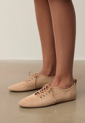 Stopy w beżowych zamszowych, sznurowanych płaskich butach z perforowanymi detalami na gładkiej beżowej podłodze na jednolitym tle.