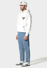 Biała bluza z kapturem z grafiką, niebieskie luźne dżinsy i białe sneakersy z czarnymi paskami. Czapka jest jasna z logo.