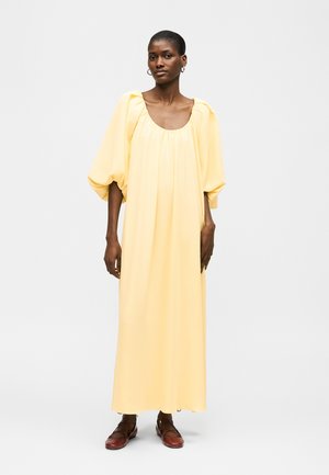 Vrouw draagt een losse, lange, lichtgele jurk met opgeblazen mouwen tot ellebooglengte en bruine platte schoenen, staand tegen een effen achtergrond.