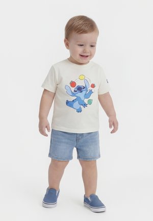 Νήπιο με λευκό μπλουζάκι που έχει ένα μπλε κινούμενο χαρακτήρα να πετάει φρούτα στον αέρα, τζιν σορτς και μπλε παπούτσια χωρίς κορδόνια, στέκεται πάνω σε λευκό φόντο.