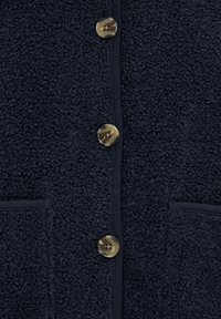 Gros plan sur un tissu polaire texturé bleu marine foncé avec trois grands boutons en écaille de tortue et vue partielle de deux poches avant.