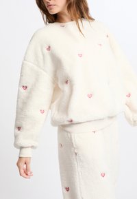 Polarowy pulower w kolorze off-white z małym haftem w kształcie czerwonego serca. Posiada luźny krój, szerokie rękawy oraz ściągacz u dołu i przy mankietach.