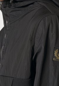Chaqueta negra con una superficie texturizada, cierre delantero con cremallera y capucha. Presenta un parche de tela con un logo en el hombro izquierdo.