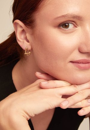 Pendientes geométricos de tono dorado con forma triangular y un lazo horizontal, llevados por una persona de piel clara y cabello rojo.