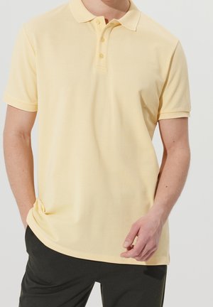 Mand iført en lysegul kortærmet poloshirt med tre knapper, kombineret med mørke bukser, stående foran en ensfarvet baggrund.