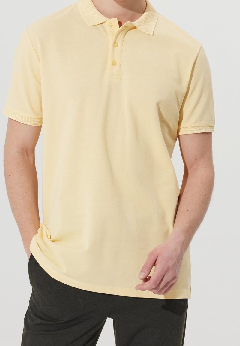 Homme portant un polo à manches courtes jaune pâle avec trois boutons, associé à un pantalon foncé, debout devant un fond uni.