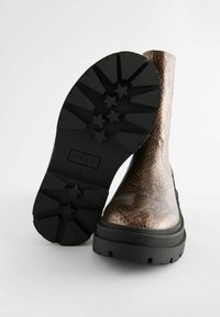 Braune Kunstschlangenleder-Ankle Boots mit einer massiven schwarzen Gummisohle, die ein sternförmiges Profil aufweist, und einer glatten, glänzenden Oberfläche im oberen Bereich.