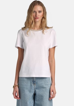 Mujer con camiseta blanca de manga corta lisa y jeans de cintura alta de color azul claro, de pie frente a un fondo liso.