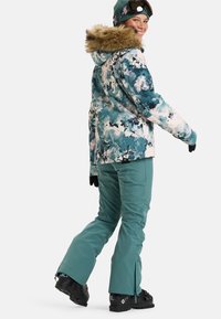 Waterdichte skijas in een blauw-wit bloemenpatroon met een nepbont capuchon. Gecombineerd met teal ski leggings, handschoenen en zwarte laarzen.