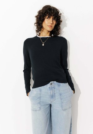 Pull noir à manches longues avec un ourlet ajusté, fabriqué en matière douce, superposé à un t-shirt à col rond blanc. Associé à un jean large bleu clair.