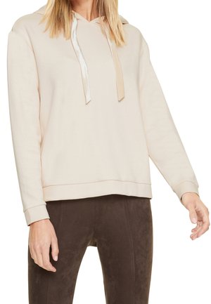 Femme portant un sweat à capuche beige avec des cordons en satin et un pantalon ajusté brun foncé, visible des épaules aux hanches, les bras détendus.