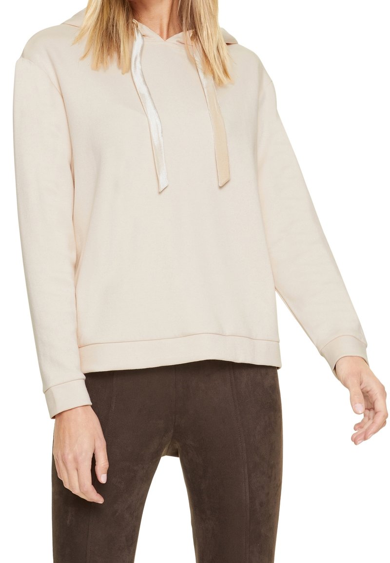 Femme portant un sweat à capuche beige avec des cordons en satin et un pantalon ajusté brun foncé, visible des épaules aux hanches, les bras détendus.