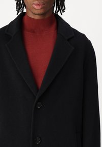 Sort uldfrakke med notch-krave og tre knapper. Båret over en rød ribbet turtleneck. Enkelt, struktureret design uden synlige mønstre.