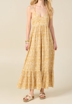 Nainen, jolla on yllään keltainen kukkakuosinen hihaton maksimekko, jossa ohuet hihnat, kerroksittain laskeutuva hame, useita rannekoruja ja beiget kävelysandaalit ilman nauhoja.