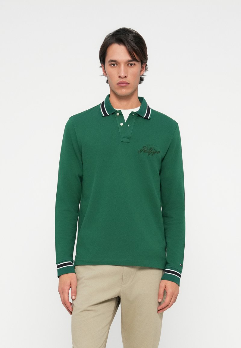 Grøn langærmet polo skjorte med en hvid indvendig krave, sort og hvid stribede detaljer og broderet logo på brystet, parret med beige bukser.