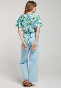 Blouse florale bleu clair à manches courtes et nouée derrière, associée à un pantalon large taille haute bleu clair et des sandales à talons brunes.