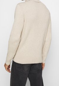 Maglione a coste beige chiaro con colletto, maniche lunghe, tessuto testurizzato e orlo leggermente curvo, abbinato a jeans scuri.