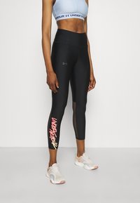 Černé legíny s vysokým pasem a síťovaným panelem, na spodní části levé nohy je vytištěný nápis "UNDER ARMOUR" v růžové a žluté barvě. Kombinováno s světlými teniskami.