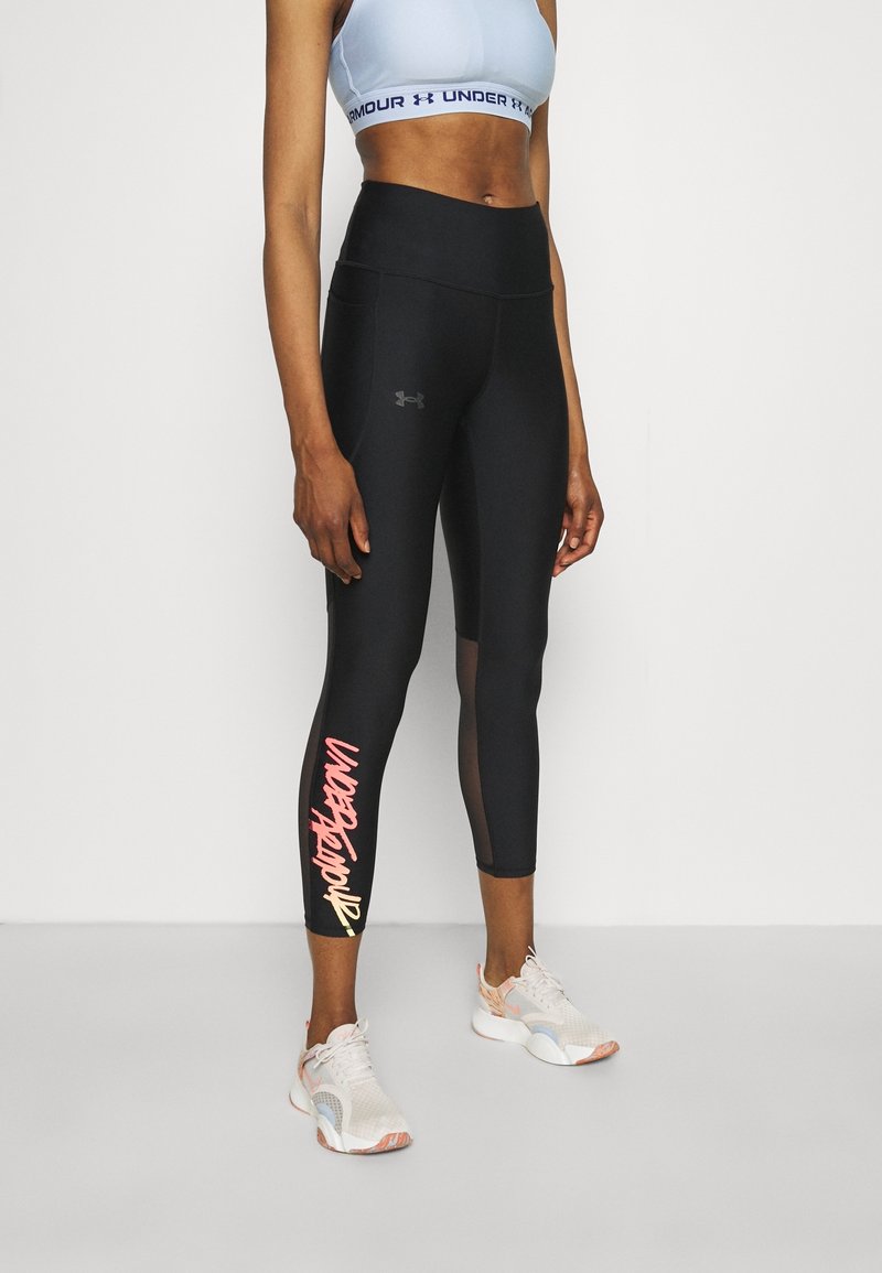 Černé legíny s vysokým pasem a síťovaným panelem, na spodní části levé nohy je vytištěný nápis "UNDER ARMOUR" v růžové a žluté barvě. Kombinováno s světlými teniskami.