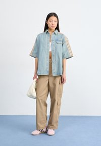 Lyseblå denim oversized skjorte med brune striber, beige cargo bukser og lyserøde sko. Modellen bærer en struktureret hvid håndtaske.