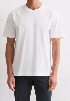 Homme portant un t-shirt blanc uni à manches courtes et un pantalon foncé, debout contre un mur clair.