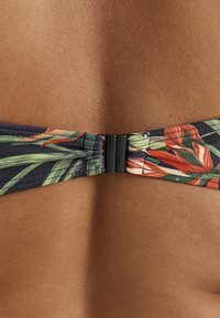 Nærbillede af en bikini-lukning, der fæstner en flerfarvet badedragt med tropisk print på en persons ryg med brun hud.