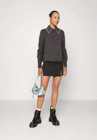 Γυναίκα με σκούρο γκρι πουλόβερ με γιακά από πέρλες, μαύρη μίνι φούστα με σκίσιμο, μαύρες στρατιωτικές μπότες, λευκές κάλτσες, κρατώντας ιριδίζουσα τσάντα.