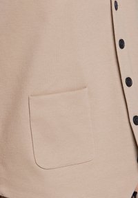 Beige tygskjorta med en texturerad yta, som har en ficka och svarta knappar på framsidan för stängning.