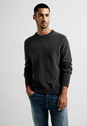 Dunkelgrauer Rundhalspullover mit gerippten Bündchen und Saum, aus einem weichen Material. Trägt dazu blaue Jeans mit den Händen in den Taschen.